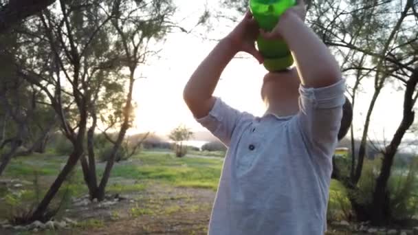 Schattige baby jongen drinkwater in het park tijdens zonsondergang — Stockvideo