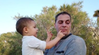 Genç beyaz bir baba oğlunu kucağında tutuyor. Bebek babasının yüzüne dokunuyor ve ikisi de komik suratlar yapıyor.