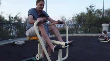 Oğlunu kucağında tutarken spor salonunda aletlerle egzersiz yapan mutlu genç bir baba. Arka planda oynayan bebek