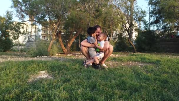 Apa a parkban guggol és megöleli a két fiát. Boldog családi koncepció — Stock videók
