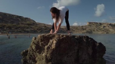 Genç beyaz kadın yoga yapıyor ve plajda kaya üzerinde esneme egzersizleri yapıyor.