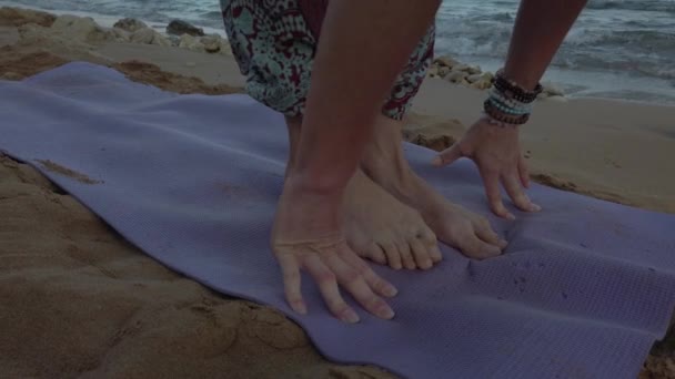 Mujer de 40 años haciendo yoga en la playa de arena por la mañana temprano. De cerca, los pies y las manos en la alfombra levantándose — Vídeos de Stock