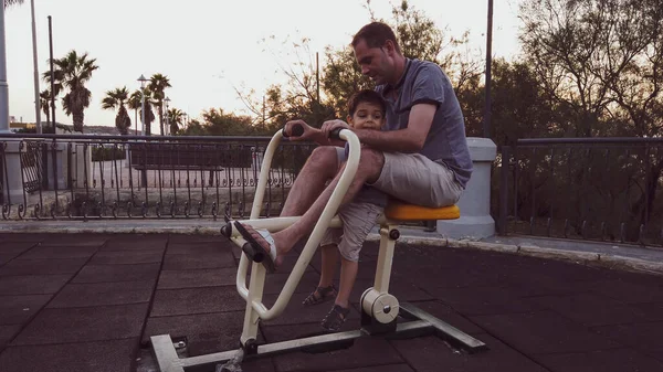 Jeune père faisant de l'exercice sur l'équipement en plein air gymnase tout en tenant son fils. Famille heureuse et concept de bien-être — Photo