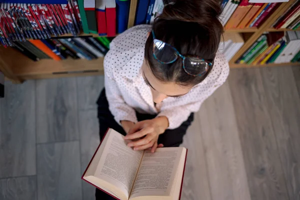 Bella donna con gli occhiali che legge un libro in libreria . — Foto Stock