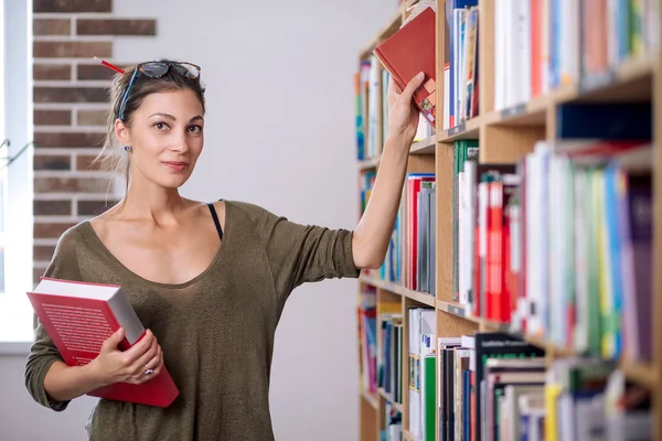 Giovane donna con gli occhiali prendere un libro dallo scaffale in una biblioteca . — Foto Stock