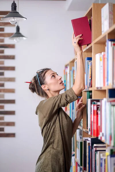 Ung kvinna som bär glasögon söker en bok i bokhyllan, jag — Stockfoto