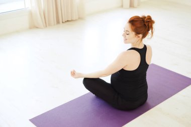 Üst kısmı geri lotus poz ayakta gülümseyen hamile kadın görmek, yüz ifadesi, diz, beyaz katta oturan ellerini dinlenme odaklanmak. Doğum öncesi yoga kavramı