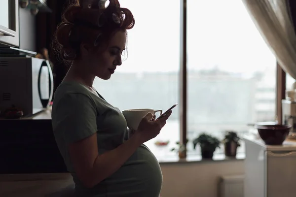 Femme enceinte utilisant un smartphone debout près de la fenêtre — Photo