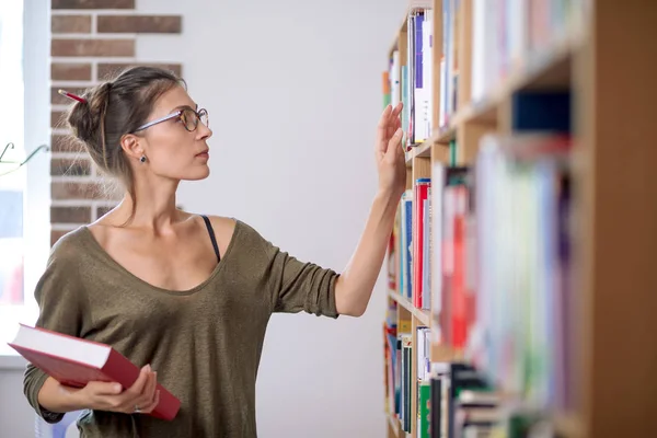 Fiatal nő visel szemüveget keres egy könyv-ra könyvespolc, én — Stock Fotó