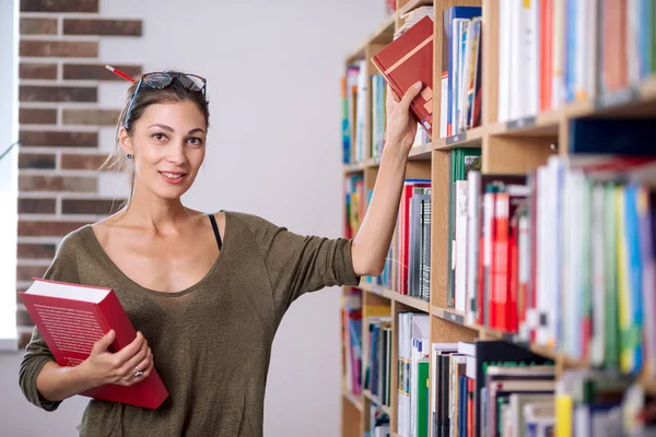 Ung kvinna bär glasögon ta en bok från hyllan i ett bibliotek. — Stockfoto