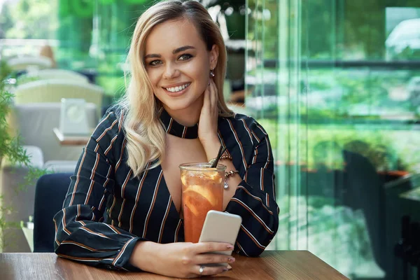 Mujer sonriente sentada en un café con una bebida fresca sosteniendo un pho — Foto de Stock