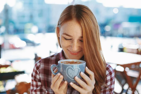 Coffee concept. Portrait of attractive young woman drinking coffee outdoors. Natural reddish girl with freckles, dressed in soft wool sweater, smiling while holding a big cup of coffee latte.