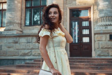 Şehir sırları. Uzakta açık havada yürürken omzunun üzerinden bakarak bir ışık elbise giyen güzel gülümseyen kadın. İyi bilinen erkeksi Yardımcısı için mutlu bir şekilde geri döndü. Şehir mimarisi.