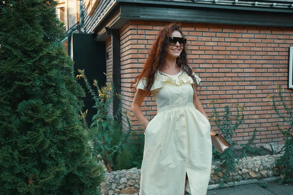 美丽的黑发年轻妇女戴墨镜 夏天黄色礼服 走在街上与房子和壶侧柏树背景 — 图库照片