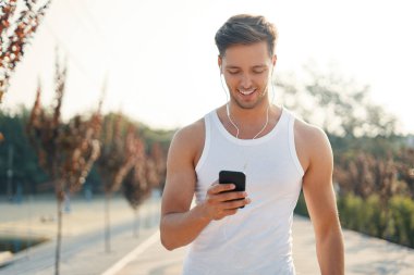 Çekici, gülümseyen, sportif genç adam onun müzik başlamak üzere kadeh çalma listesi üzerinde smartphone için ekran açık havada bakarken.