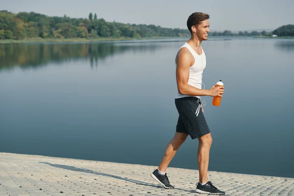 微笑和放松运动男子的肖像 手里拿着一瓶水 身着黑色运动衫的运动员穿着白色耳机享受早晨 而在附近的湖边慢跑 — 图库照片