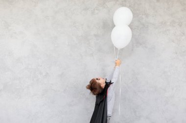 Cadılar Bayramı vampir siyah pelerin giyen, gülüyor ise, gri dokulu bir arka plan üzerinde siyah iri gözlü el 2 beyaz balon hayaletler germe çocuk profili. Kopyalama alanı.