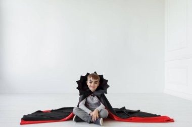 Gülümseyen oğlan vampir makyaj ile kırmızı, siyah pelerin gibi yaka, arka plan üzerinde beyaz hayaletler ile hafif bir alanda sopası ile çapraz ayaklı oturuyor. Cadılar Bayramı ruh, kopya alanı oynama.