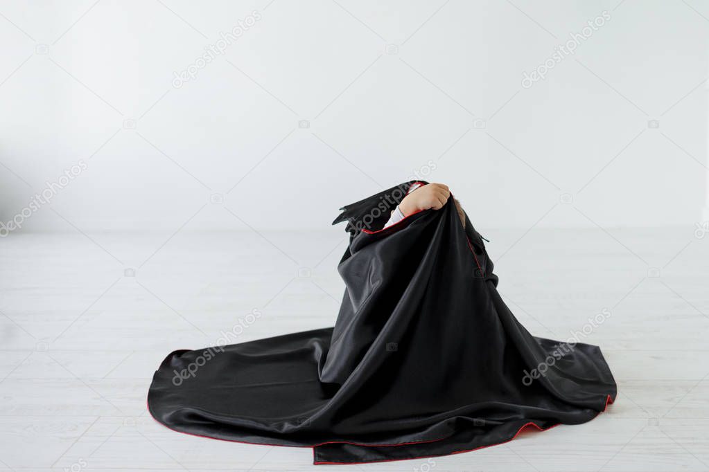 Kids playing concept. Playing boy hiding himself with a black cloak imagining he is a sleeping vampire or invisible man. Halloween mood, creative kids. Poster, promotions, advertising friendly.