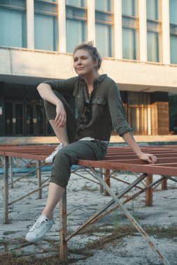 Metal konstrüksiyon ile arka binada oturan bir casual sarışın kız portresi. Genç modern kadın şehirde, açık havada poz. Kentsel yaşam