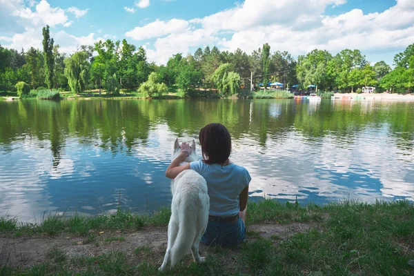 Best friends, woman hugging a dog, resting while enjoying the amazing view of landscape, lake reflecting the sky and trees. Back view.