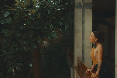 Otel balkonda duran, egzotik ağaçlarıyla çevrili bir tatil zevk dışarı arayan çok şehvetli genç kadın. Dinlenme ve rahatlama kavramı.