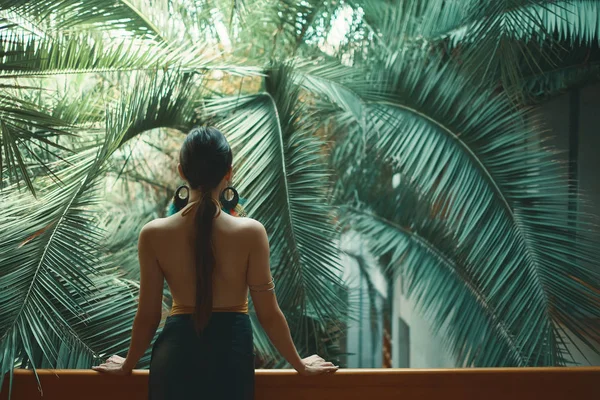 Back view fit exotic woman, bare back, watching huge palm tree leaves while resting on the balcony, arranging her hair, extending her hands up. Resort and vacations promotion. Beautiful life concept.