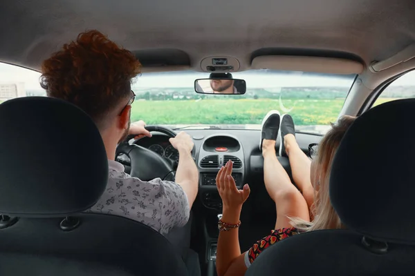 Happy couple enjoying on a long drive in a car. Friends driving on road trip on summer day. Caucasian young man driving a car and joyful woman with her arms raised.