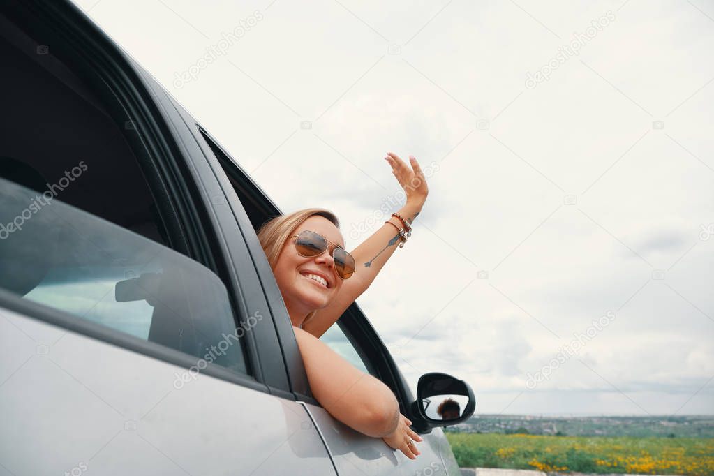 Happy  young woman enjoying the views on a road trip while looking through the car window, saying hello waving her hand.