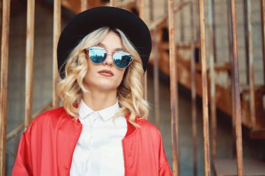 Güzel ve şık bir genç kadın paslı metal merdivenlerde, şık giysiler, kırmızı ceket, beyaz gömlek, siyah şapka ve güneş gözlüğü takıyor teşkil etmektedir. Pop kültürü. Moda hipster portre.