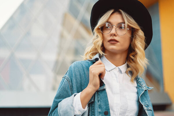 Close-up portrait of a cheerful white woman in glasses touching her jacket collar on urban background. Photo of fashionable girl with beautiful blonde hair posing for the camera.
