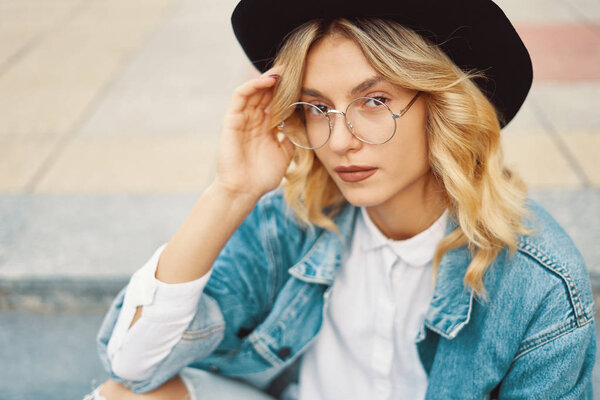 Close-up portrait of a cheerful white woman in glasses touching her hat on urban background. Photo of fashionable girl with beautiful blonde hair posing for the camera.
