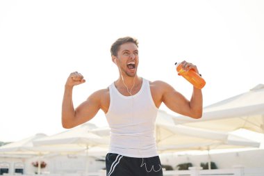 Atletik kaslı adam duygu zafer ve bir şişe su tutarak başarı sahiptir. Duygu, güç, jest - zaferini kutluyor ve ile güneş ışığı arka planda çığlık genç adam.