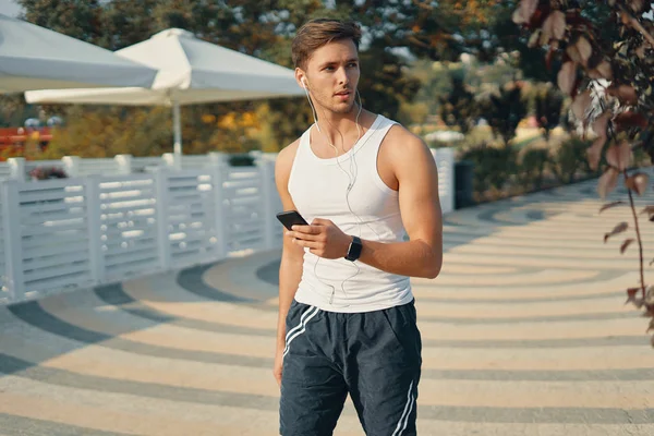 Young, handsome runner with smart phone, watch and earphones outside in sunny autumn. Sport and technology concept.