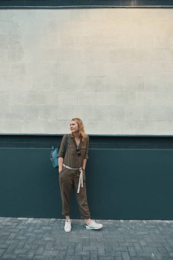 Uzun sarı saçlı bir hippi kız, bir şık romper ve bir sırt çantası takmış bir yana duran bir duvar arka planda bir sokakta arıyor. Yatay yukarı alay etmek. Metin veya tasarım için boş alan.