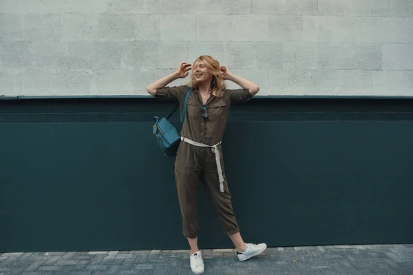 一个时髦的女孩 长着金色的头发 穿着时髦的爬衣和背包 一边站在墙上的背景上的街道上 水平模拟 文本或设计的空白空间 — 图库照片
