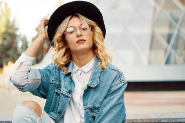 Yakın çekim gözlük şapkasını kentsel arka plan üzerinde dokunmadan neşeli beyaz bir kadın portresi. Kamera için poz güzel sarışın moda kız fotoğrafı.