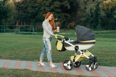 Küçük çocuğunu onun bebek arabası rahatlatıcı ile sıcak bir çay içme park yürüyüş zevk içinde sabah, Genç bir anne.