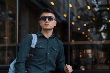 Modern genç erkek güneş gözlüğü ve ışıklar arka planda karanlık pencere karşı duran sırt çantası