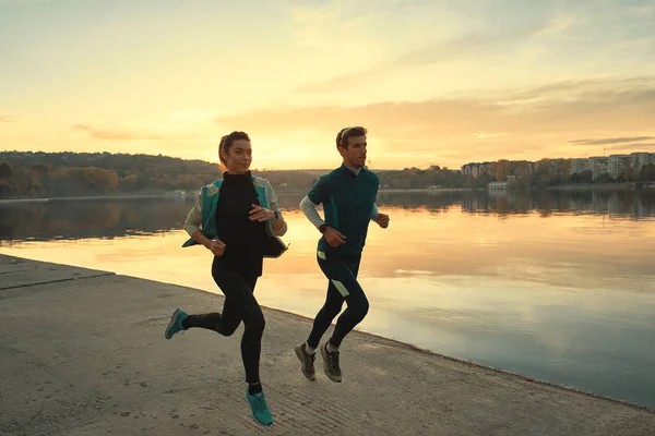 在日出的时候 激励着一对跑步者在湖面上跑步 穿着运动服的年轻男女 一起跑步 户外运动训练人员 — 图库照片