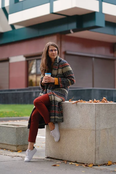 屋外コーヒーを飲みながらコートでスタイリッシュな若い女性 現代都市の服装と休憩をしながら街の通りに座っているメガネのカジュアルな女の子 — ストック写真