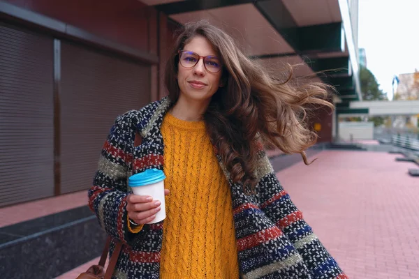 Urban lifestyle and coffee on the go. Stylish and  young woman in coat drinking coffee outdoors. Casual girl in modern urban outfit and glasses walking city streets.