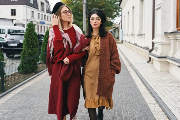 Carefree fashionable women models in elegant autumn clothes and glasses. Young fashion girls in stylish woven wool clothes and hats over urban city background, autumn portrait.