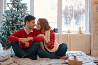 Oturma ile dekore edilmiş ağaç bir yatakta çapraz bacakları ve arka plan üzerinde pencere ile gülümseyen kız gözlerini için arayan genç adam. Noel zevk mutlu sevgi dolu Çift.