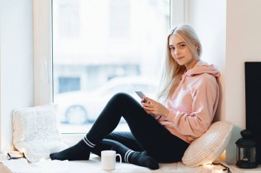 Bir fincan kahve, çay ve ellerde smartphone ile pencere eşiğinde oturan güzel kız. O uzun sarı saçlı, gülümseme ve seyir onun telefon vardır. Pembe, soluk parka giyiyor.