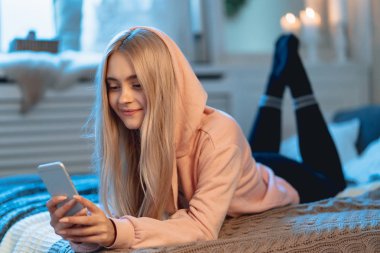 Sarışın kız pembe parka ve arkadaşları ile sohbet ederken evde, yatakta yatıyordu bir akıllı telefon kullanarak siyah tozluk gülümseyen