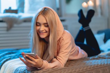 Sarışın kız pembe parka ve arkadaşları ile sohbet ederken evde, yatakta yatıyordu bir akıllı telefon kullanarak siyah tozluk gülümseyen