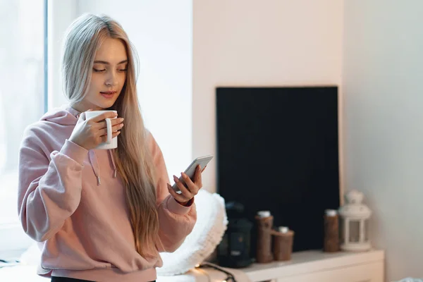 Csinos Lány Ablakpárkányon Egy Csésze Kávé Tea Smartphone Kezében Van — Stock Fotó