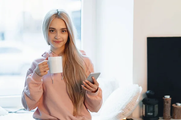 Csinos Lány Ablakpárkányon Egy Csésze Kávé Tea Smartphone Kezében Van — Stock Fotó