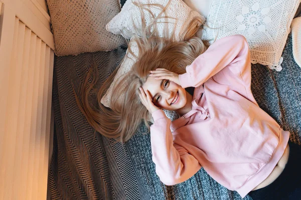Beautiful smiling girl, with long blond hair, enjoying happiness, she cant belive its true. Portrait gorgeous young woman lying on the bad with pillows in comfy pink pale parka looking at camera.